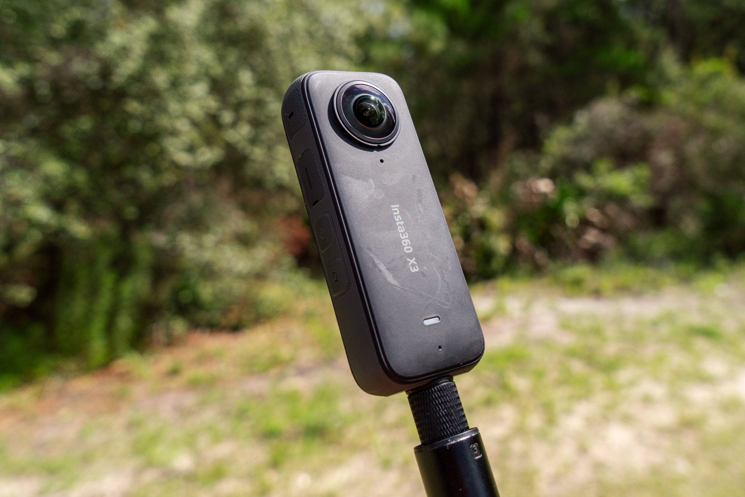 The Insta360 X3 on a selfie stick in front of woods.